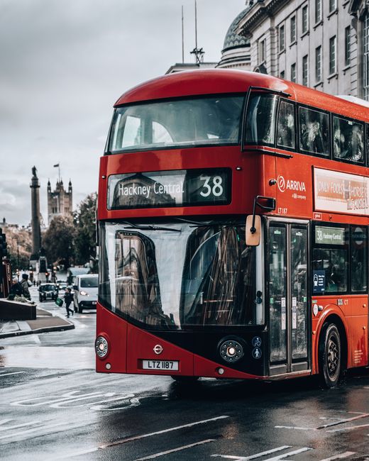 A red double-decker bus on a wet street in Shoreditch, with reflections on the shiny surface and nearby buildings visible through the large front windows. The bus displays the route number 38 and is a
